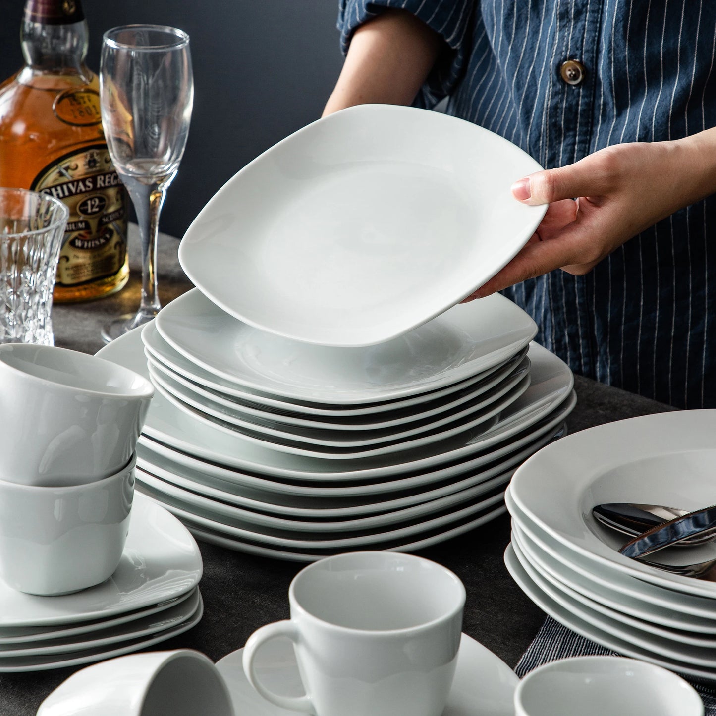 Stacks of white ceramic plates and cups on a table with a bottle of Shivas Keval in the background.