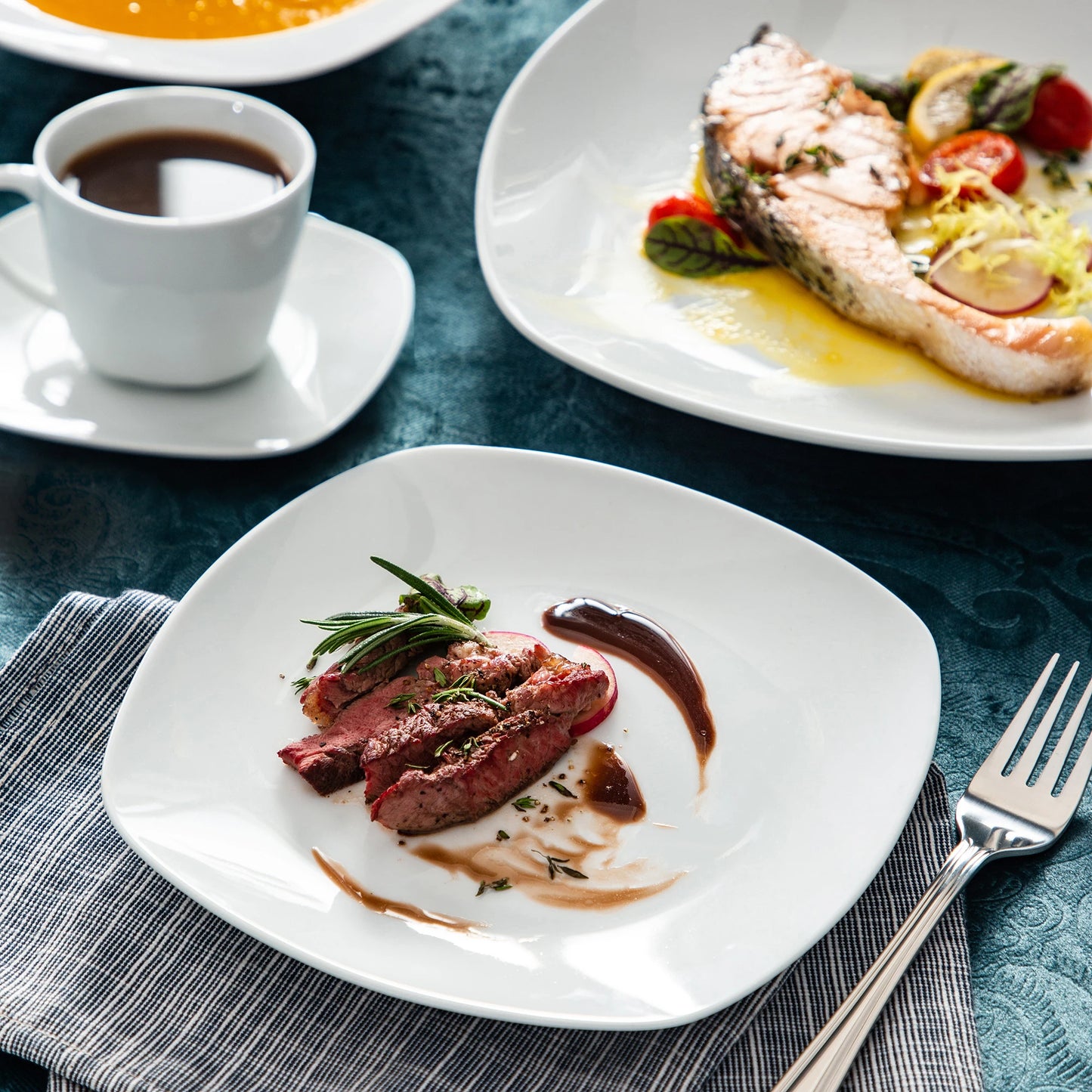 Dinner setting with a plate of meat, a cup of coffee, and a side dish on a blue tablecloth.